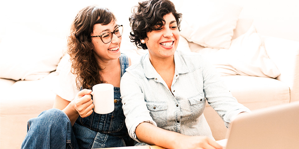 Image of two smiling women sitting on a couch and looking at a laptop screen.