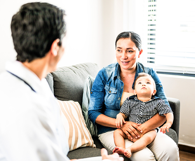 A mother holding her baby on her lap sit on a couch talking to a medical professional.