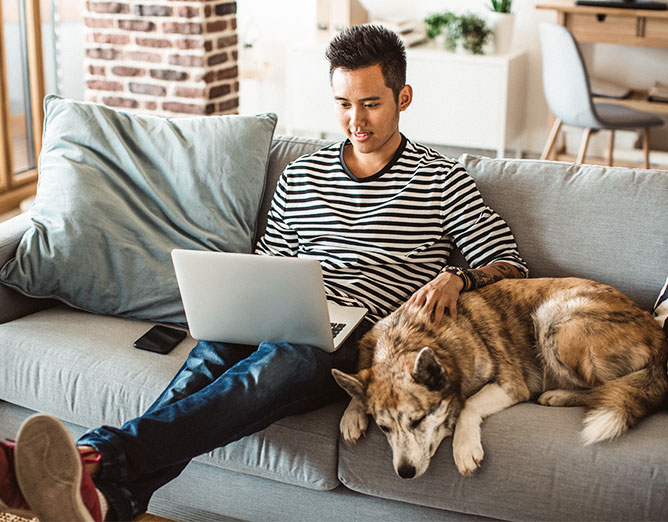 Adult male using a laptop computer while sitting next to a dog on a couch